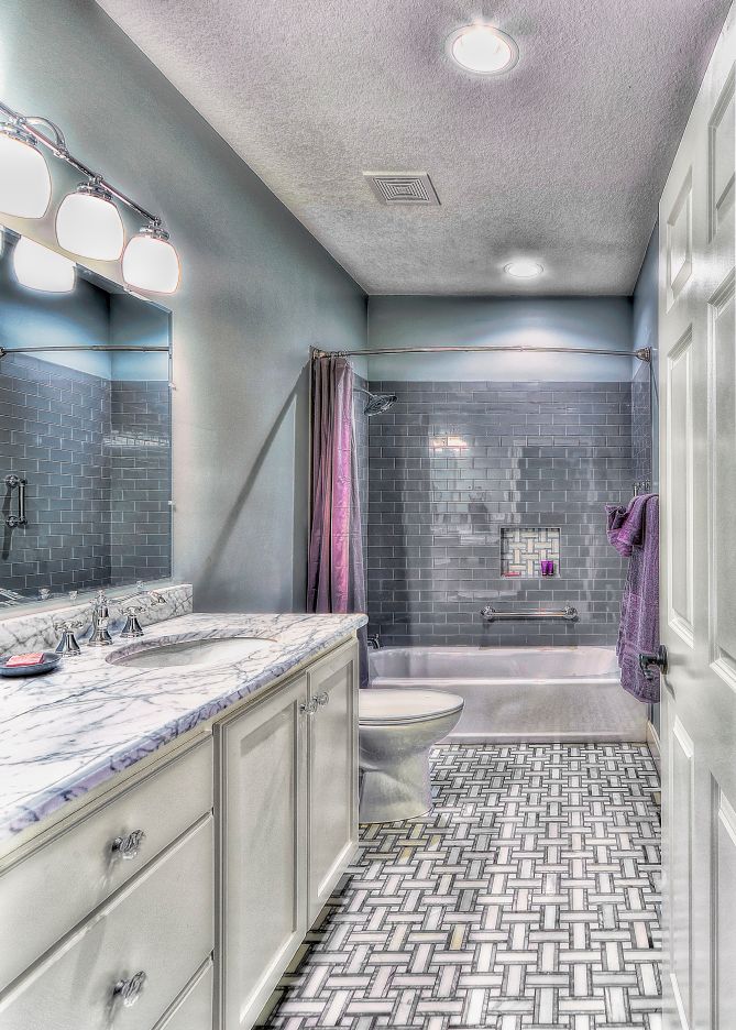 A bathroom with gray walls, white cabinets, a marble countertop, patterned tile floors, and a tiled shower-tub combo.