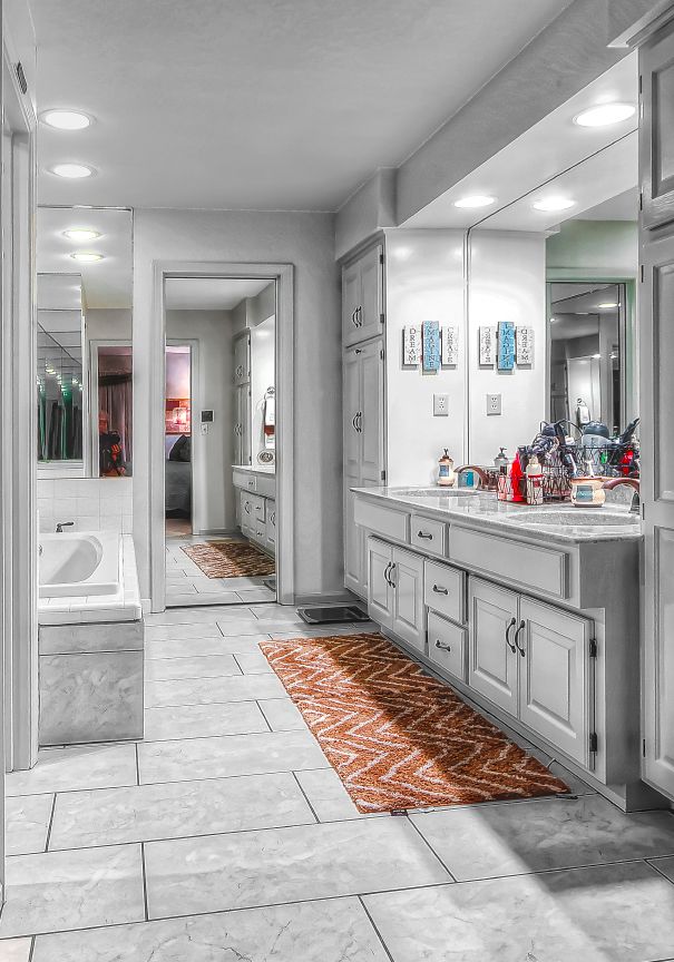 A bright, neutral-toned bathroom with a double vanity, large mirror, white cabinets, a soaking tub, and a patterned rug.
