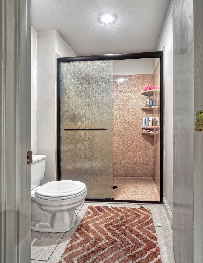 A small bathroom featuring a toilet next to a glass-doored shower with a pink tiled interior and a chevron rug on the floor.