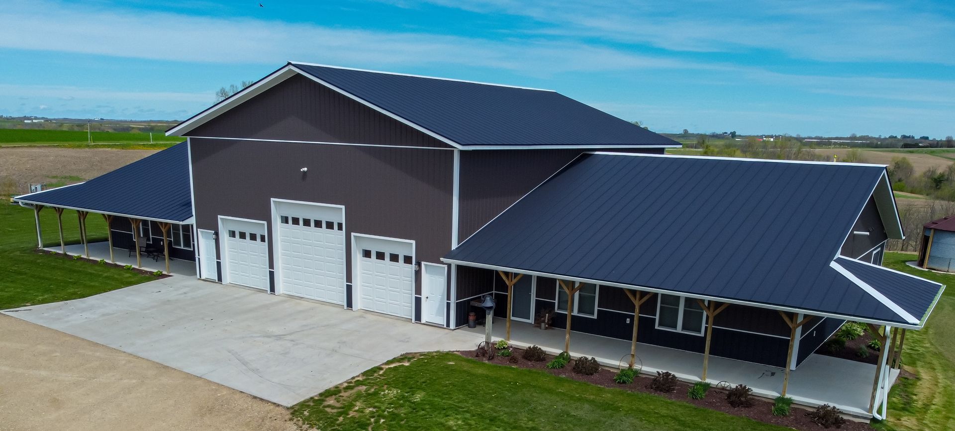 Aerial view of a dark brown barn-style building with a metal roof, three garage doors, and a covered porch in a field.