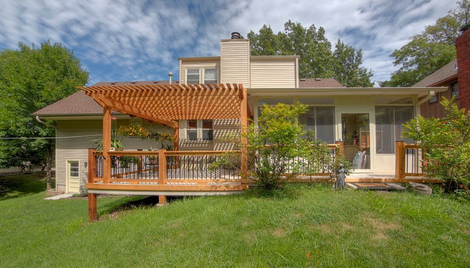 A wooden deck with a pergola attached to the back of a two-story beige house, overlooking a grassy backyard.