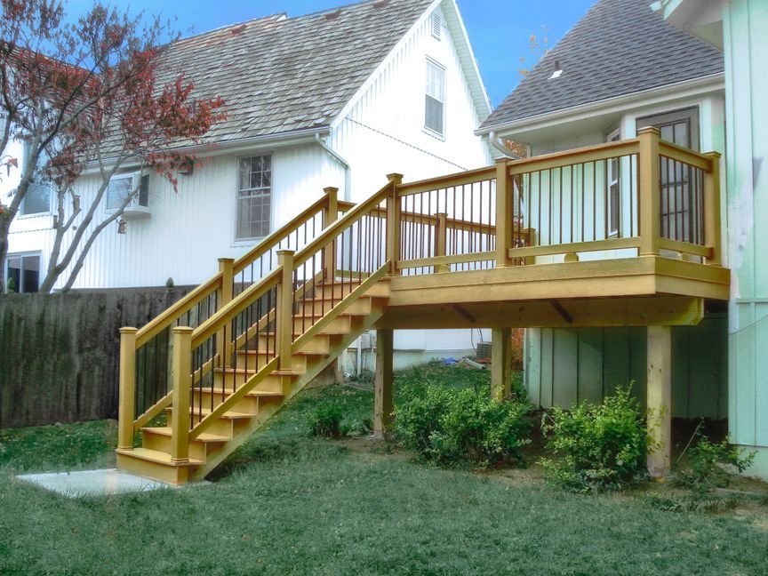 A wooden deck with a staircase leading down to a grassy yard next to a two-story residential house.