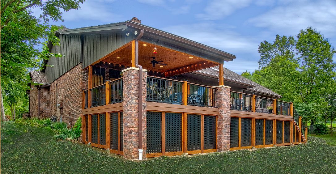 A rustic stone-and-timber house with a large, elevated deck, covered patio, and landscaped lawn on a sunny day.