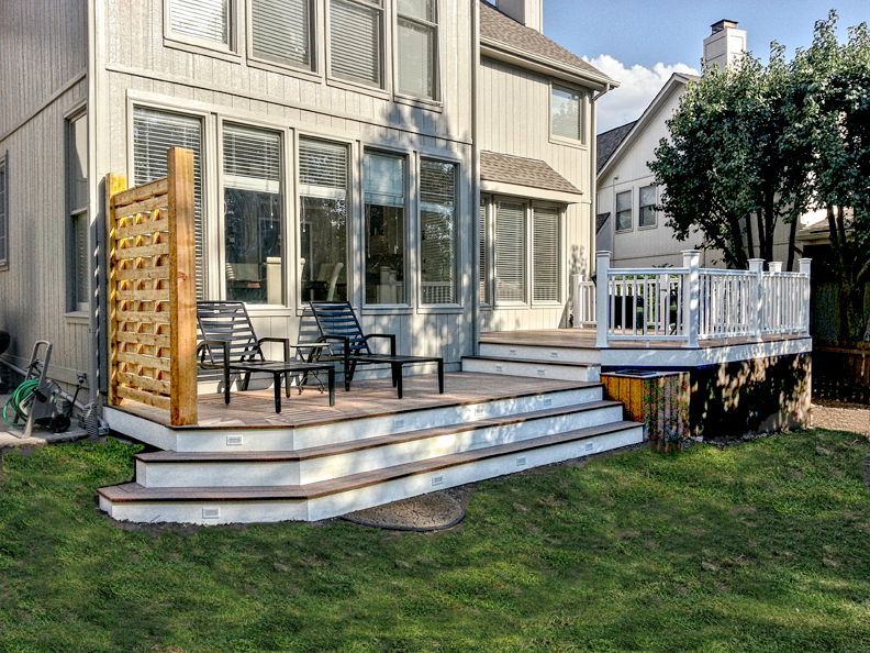 A light gray house with a two-tiered wooden deck, white railing, privacy screen, and two outdoor chairs on a lawn.