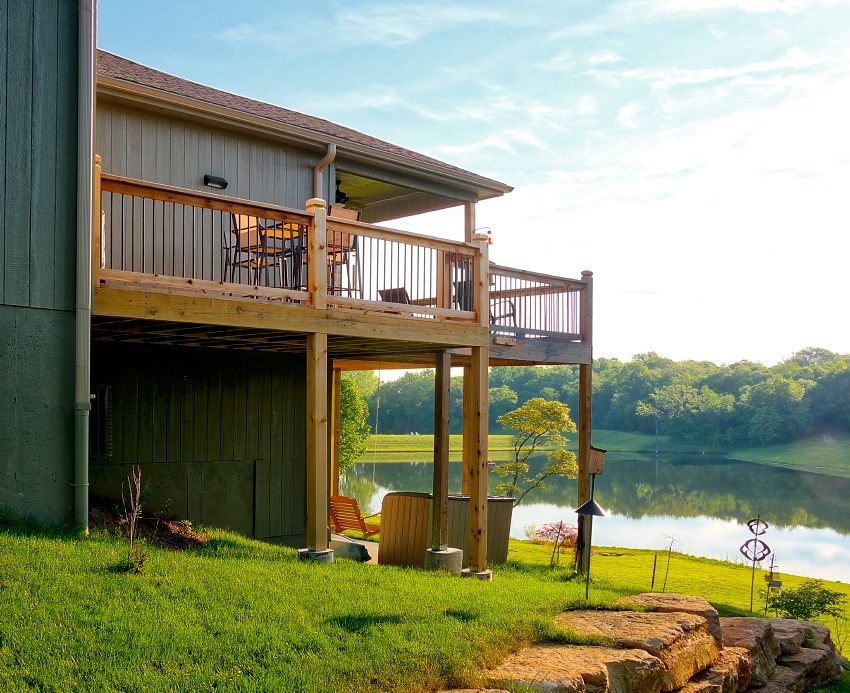 A house with a large wooden deck overlooking a lake and grassy hill under a bright, sunny sky.