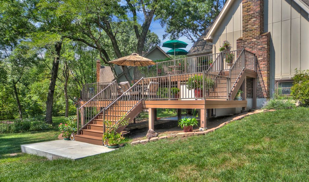 A wooden deck with a staircase and a patio umbrella extends from a house into a grassy, tree-filled backyard.
