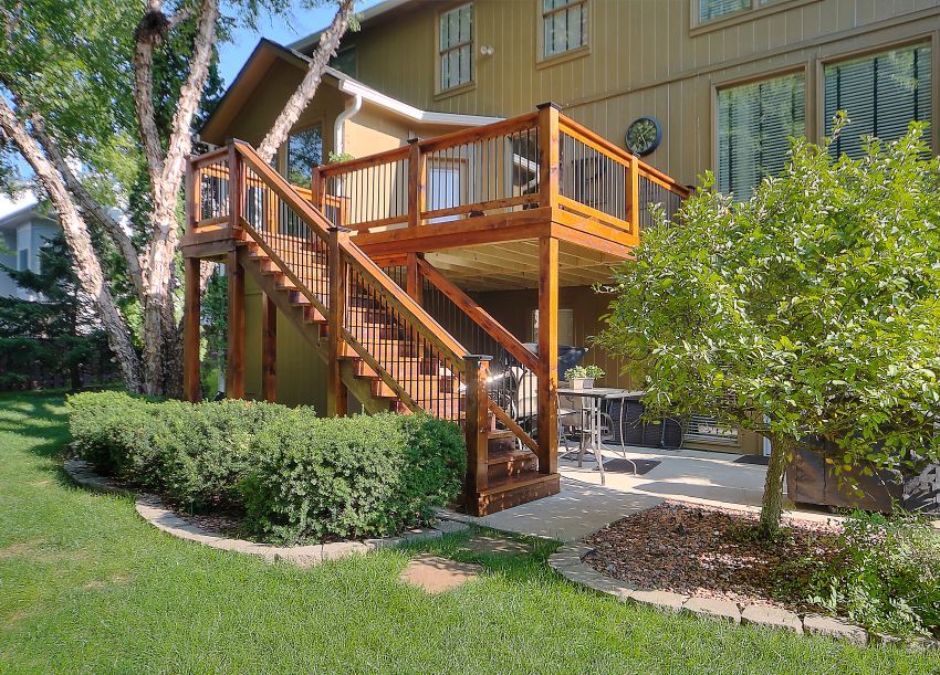 A wooden deck with a staircase leading down to a paved patio area in a backyard with trees and green lawn.