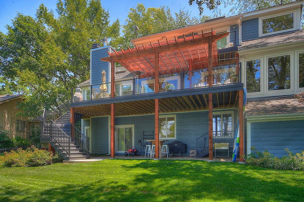 A blue two-story house with a wooden pergola over a deck, surrounded by a lush green lawn and trees.
