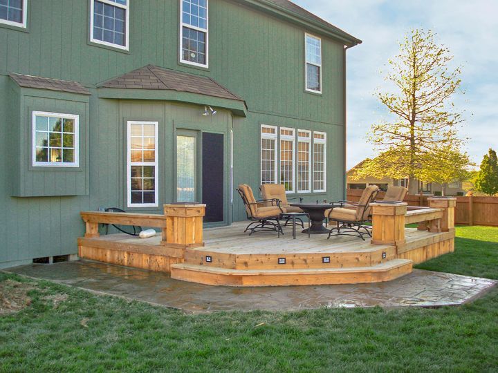 A wooden deck with a table and chairs set against the back of a green, two-story house with large windows.