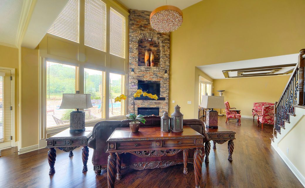 A sunlit living room with yellow walls, a stone fireplace, dark wood furniture, and large windows looking outdoors.
