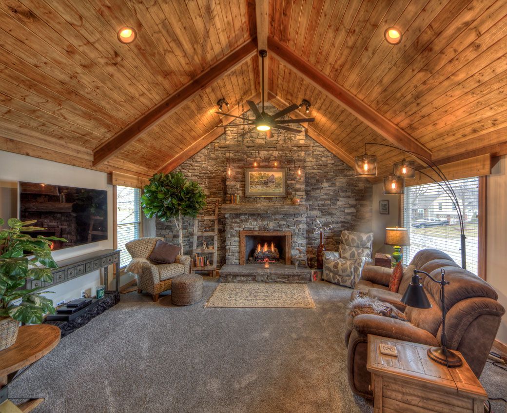 A rustic living room with a stone fireplace, wood-paneled vaulted ceiling, comfortable furniture, and a television.