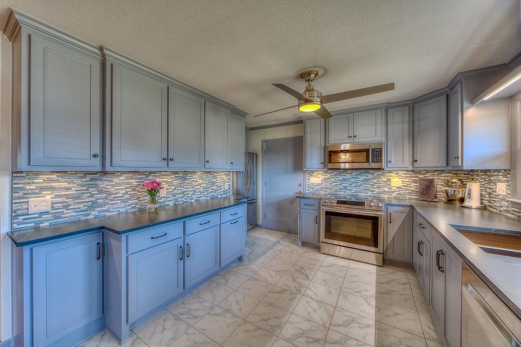 A bright kitchen features blue cabinetry, a patterned backsplash, dark countertops, and a ceiling fan.