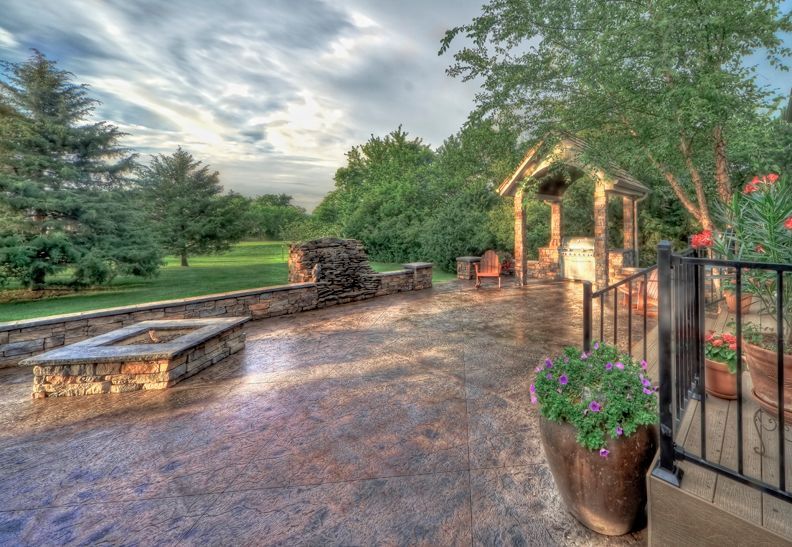 A stone patio with a fire pit, outdoor kitchen, and landscaping under a cloudy sky.
