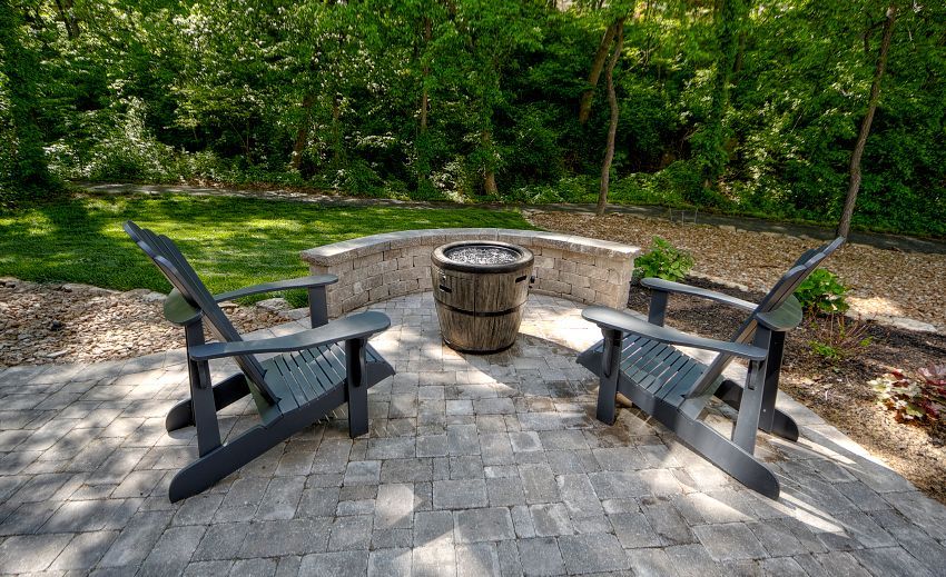 Two gray Adirondack chairs face a stone fire pit on a paver patio, set against a backdrop of trees and lawn.