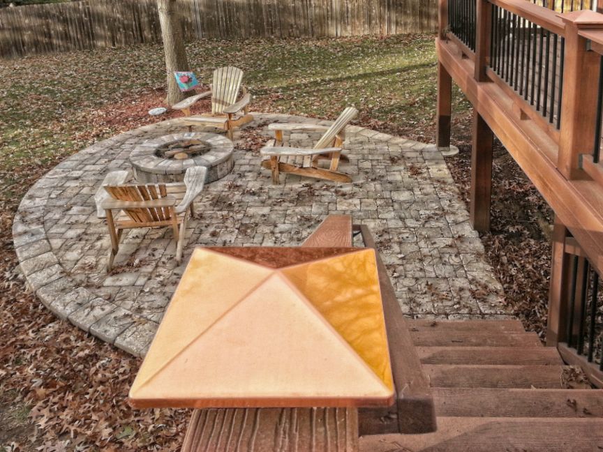 A wooden deck staircase descends toward a circular stone patio with a fire pit and several wooden chairs in a backyard.