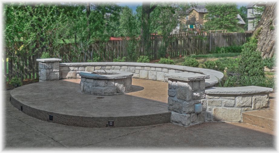 A curved outdoor patio features a stone fire pit, a raised seating wall, and pillars, set against a wooden fence and garden.