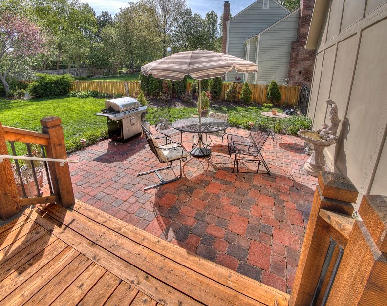 A sunny backyard patio featuring red brick pavers, a dining table set with an umbrella, a grill, and a wooden deck.