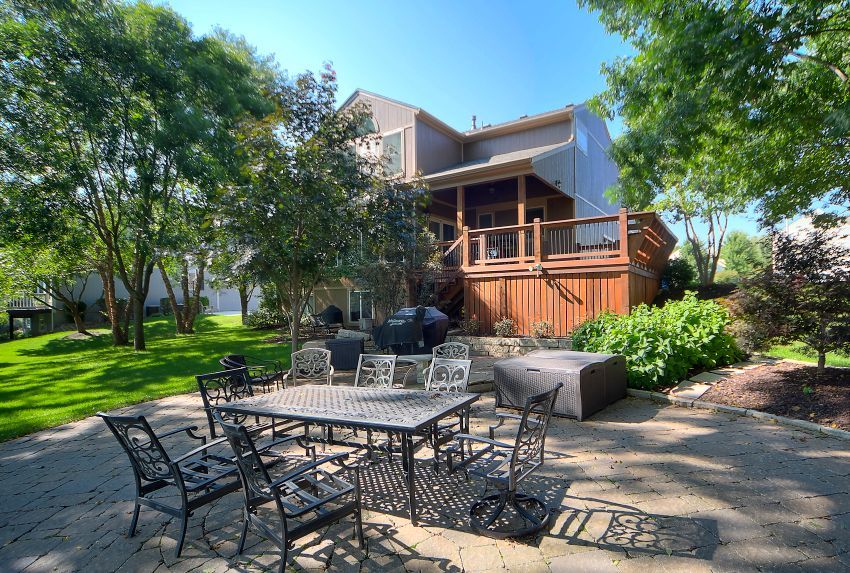 A patio with a dark metal dining set overlooks a green lawn and trees, with a two-story house and wooden deck in the back.