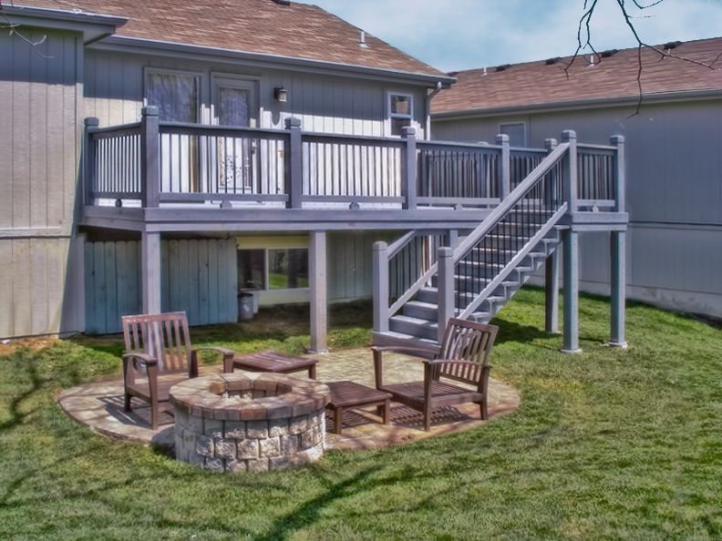 A gray wooden deck with stairs leads to a backyard stone patio featuring a fire pit and two wooden chairs.