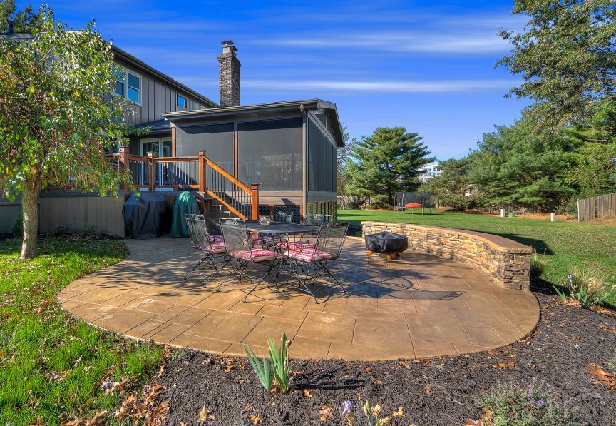 A backyard patio with a stone retaining wall, outdoor table, chairs, and a screened-in porch attached to a house.