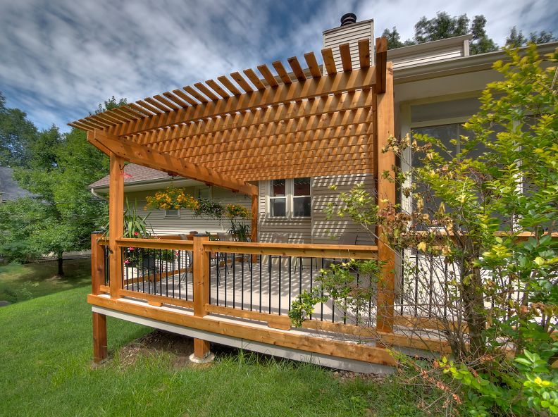 A wooden pergola covers a backyard deck featuring black metal railings, set against a house exterior on a grassy lawn.