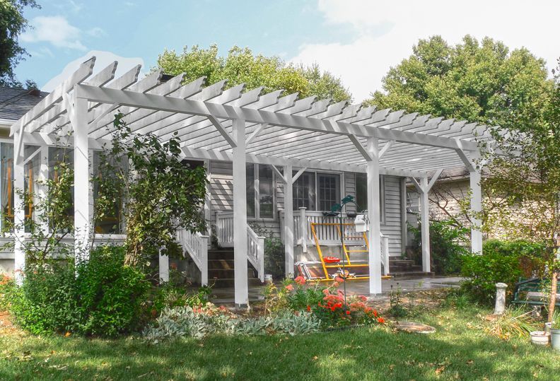 A large, white wooden pergola attached to the back of a house, overlooking a grassy yard with plants and a swing.