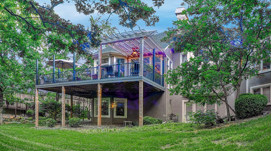 A multi-level home with a large wooden deck featuring a pergola, string lights, and patio furniture, surrounded by trees.