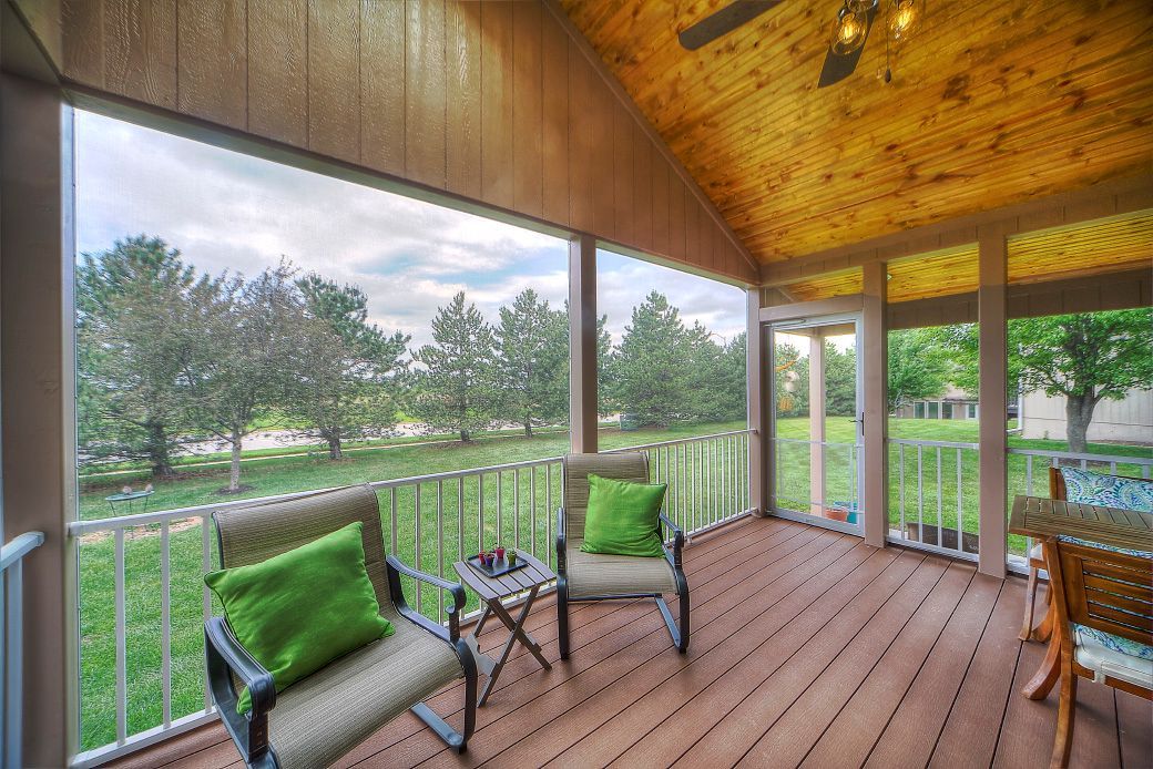 A screened-in porch with wood ceiling, two chairs with bright green pillows, a side table, and a view of a grassy yard.