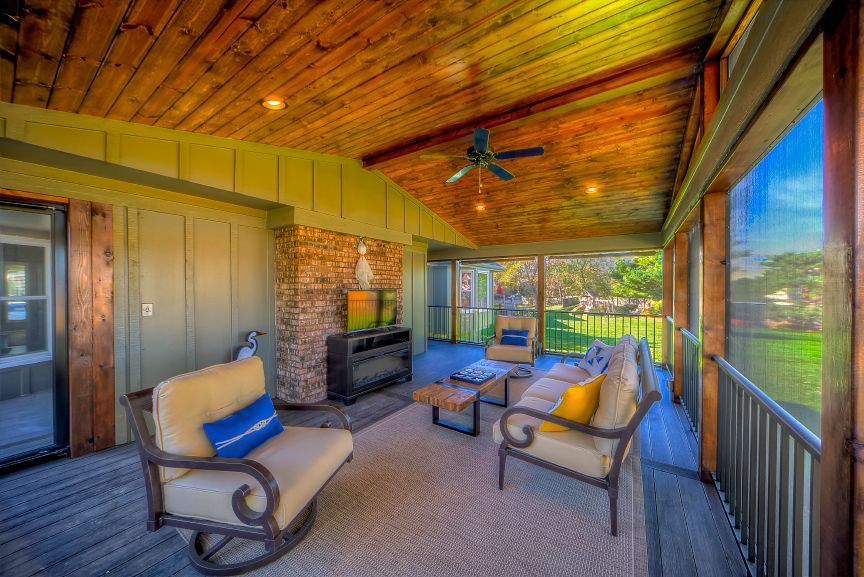 A screened-in porch with a wooden ceiling, stone fireplace, outdoor furniture, and views of a green lawn.