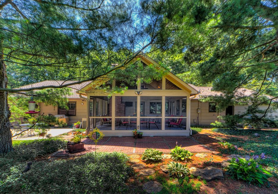 A brick patio leads to a screened-in porch at the back of a beige house, surrounded by lush trees and green landscaping.