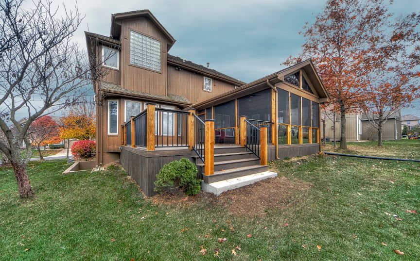 A two-story brown house with a large wooden deck and attached screened-in porch, situated on a lawn in autumn.