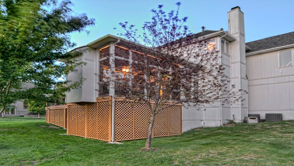 A multi-story beige house features an elevated screen porch with wooden lattice skirting, surrounded by a grassy lawn.