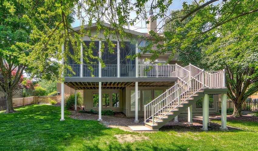A two-story tan house with a screened-in upper deck and stairs leading down to a green lawn surrounded by trees.