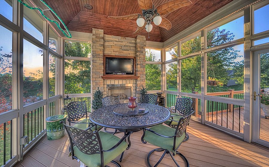 A screened-in porch with a wooden ceiling, stone fireplace, flat-screen TV, and a patio dining set with green cushions.