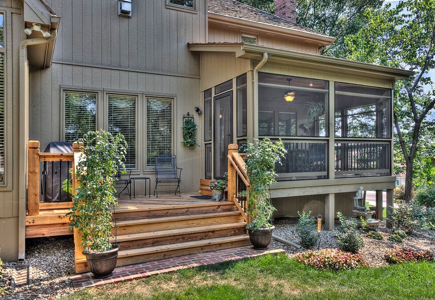 A tan wooden deck and screened-in porch attached to a tan house with outdoor plants and stairs leading to the lawn.