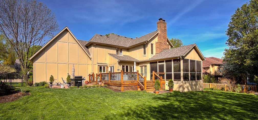 A two-story stucco house with a wooden deck and a screened-in porch on a sunny, green lawn under a clear blue sky.