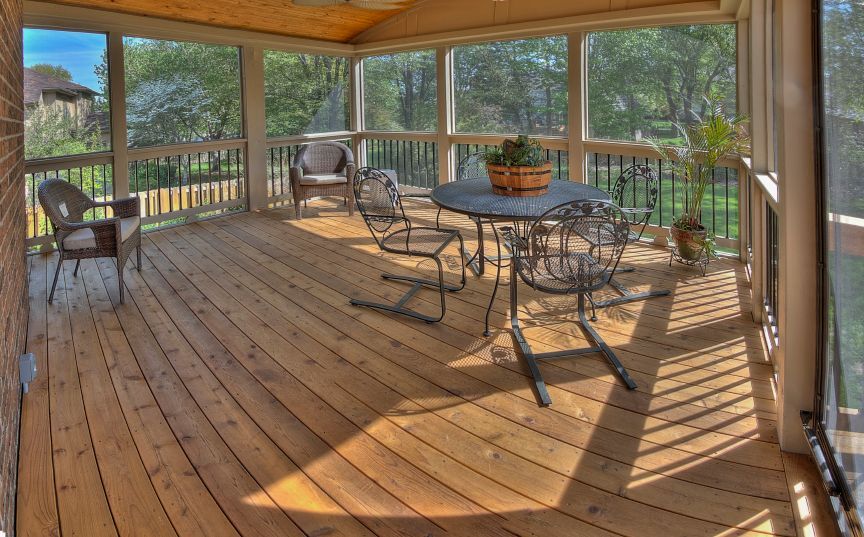 A screened-in porch with natural wood flooring, a central round table with three metal chairs, and one armchair.