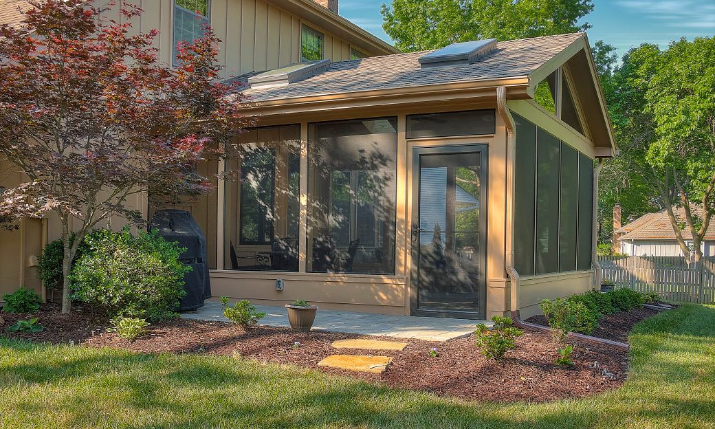 A beige, modern sunroom addition with screen walls and a dark-framed door attached to the back of a tan suburban house.