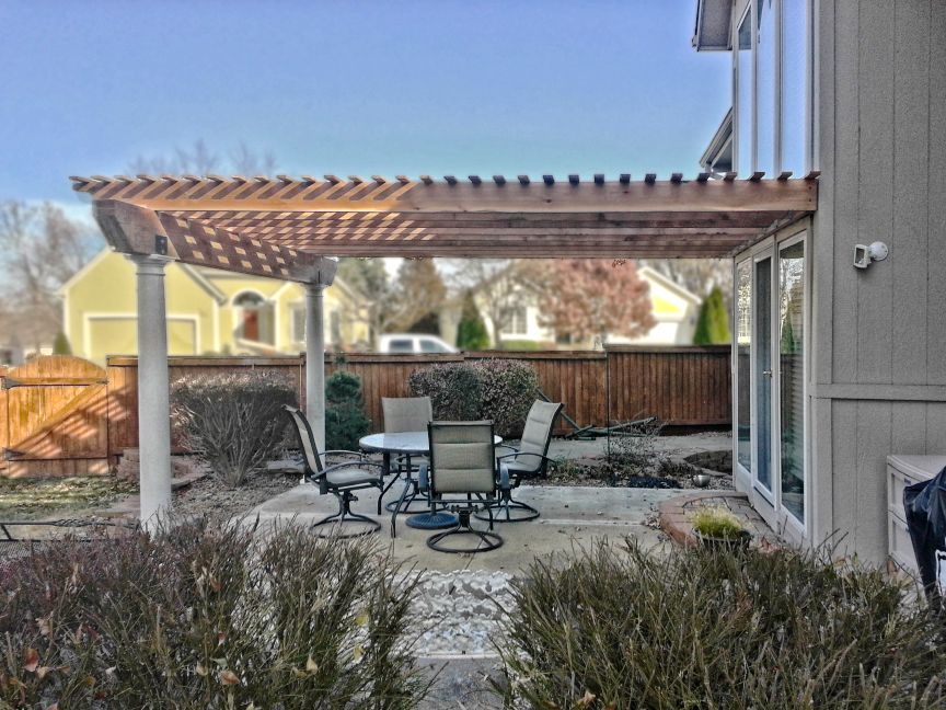 A patio featuring a wooden pergola, dining set, and surrounding bushes against the side of a house.