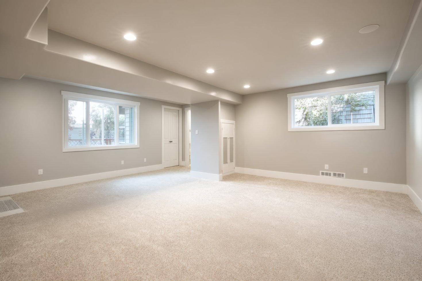 A spacious, empty basement with beige carpeting, light grey walls, recessed ceiling lights, and two windows.