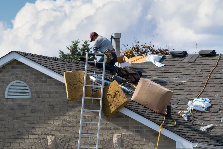 A worker in a cap climbs a ladder onto a residential roof covered with shingles, insulation, and cardboard debris.