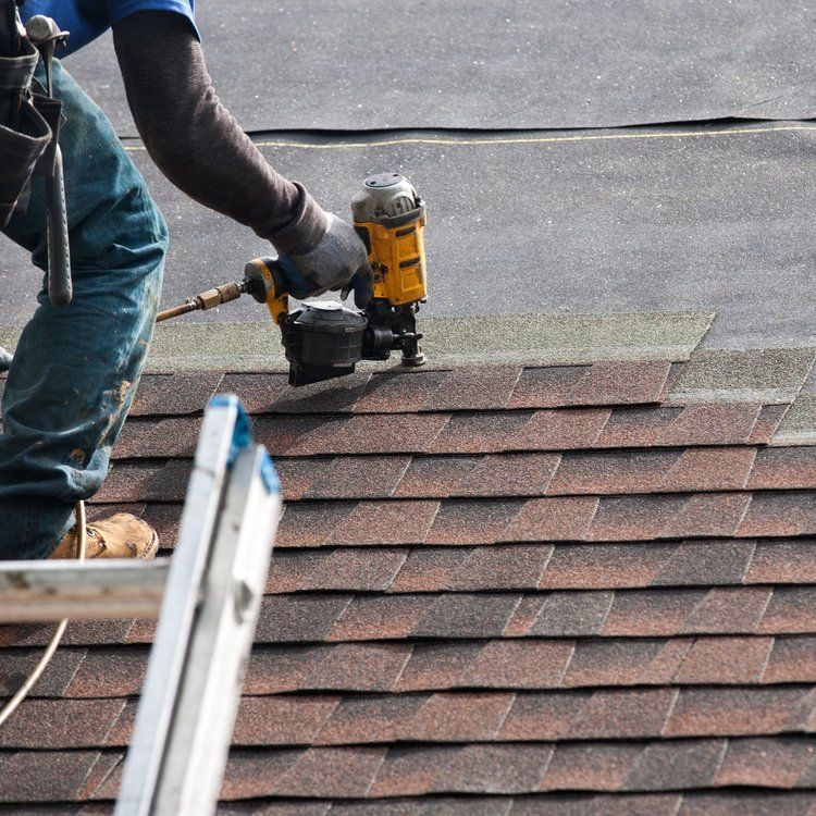 Worker using a nail gun to install shingles on a roof, with a ladder nearby.