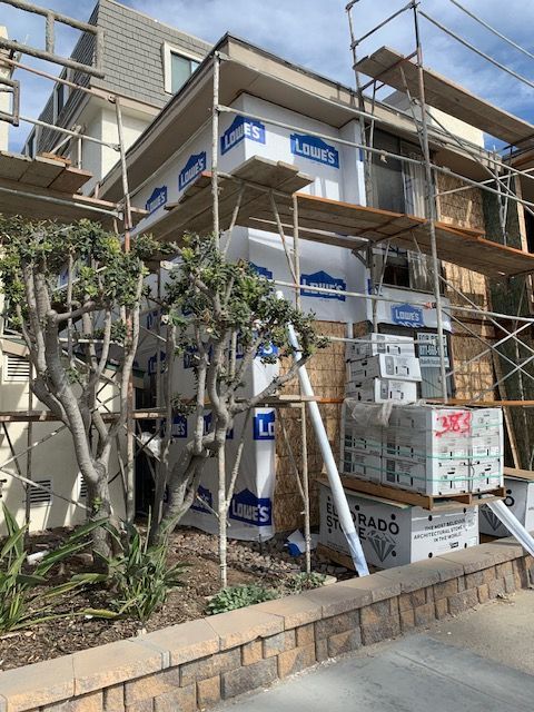 White stucco building with scaffolding and blue signs behind trees on a sloped sidewalk
