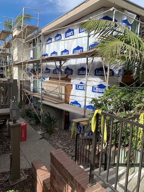 Scaffolded building with blue and white wrapped exterior, viewed from a walkway beside plants and a fence