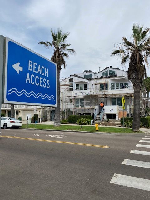 Blue “Beach Access” sign beside a street and crosswalk near palm trees and a white building.