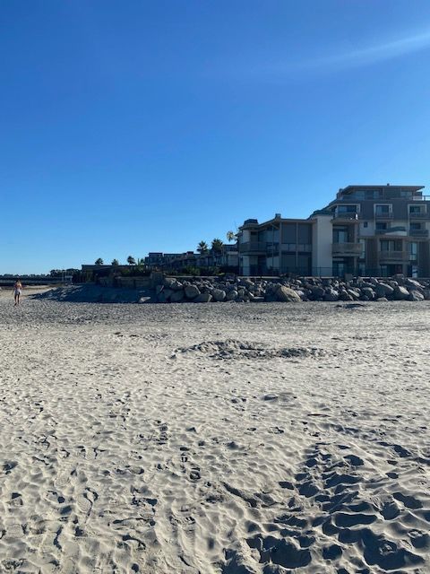 Sandy beach under a clear blue sky, with seaside buildings and rocks in the distance