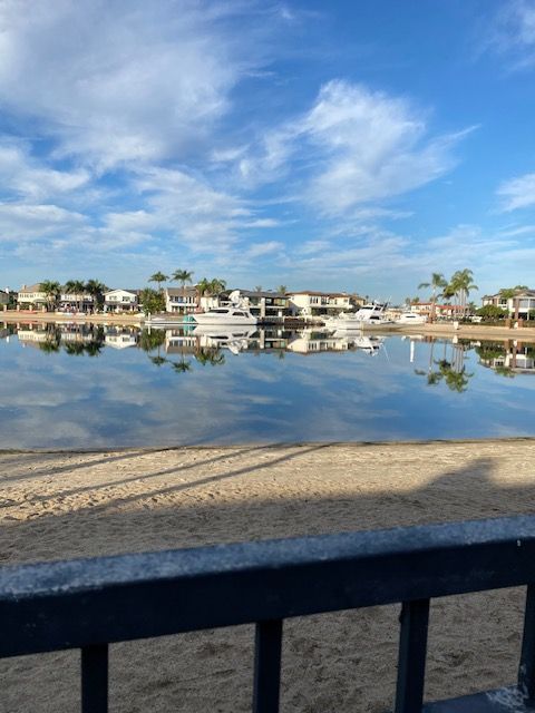Calm waterfront with houses reflected in still water under a blue sky, viewed from a sandy shore.