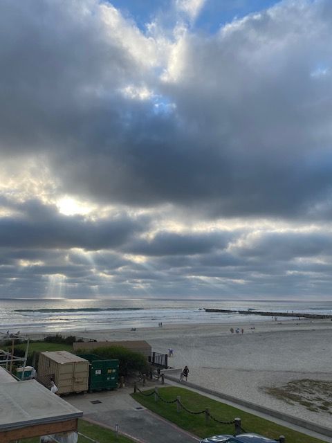 Cloudy beach scene with low tide, people in the distance, and buildings beside a grassy walkway