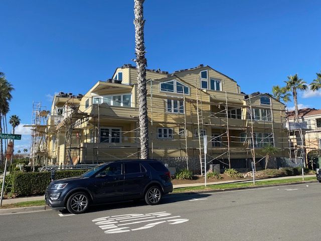 Yellow apartment building under construction with scaffolding and a black SUV parked in front under a blue sky