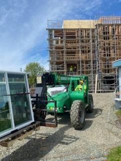 Green telehandler lifting a window frame at a construction site with a wooden building under scaffolding